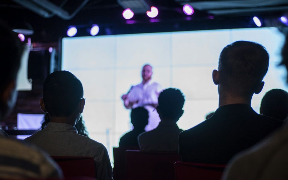 An audience watching a speaker present on a large screen